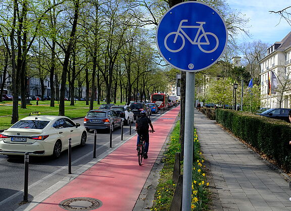 Ein geschützter Radfahrstreifen mit Roteinfärbung. Radwegschild und einer Radfahrerin. 