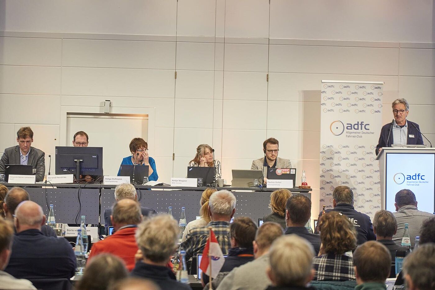 45. ADFC-Bundeshauptversammlung 2025 Mehrere Personen sitzen auf einem Podium vor Laptops, während rechts Christian Tänzler am Pult zu einem vollen Saal spricht.